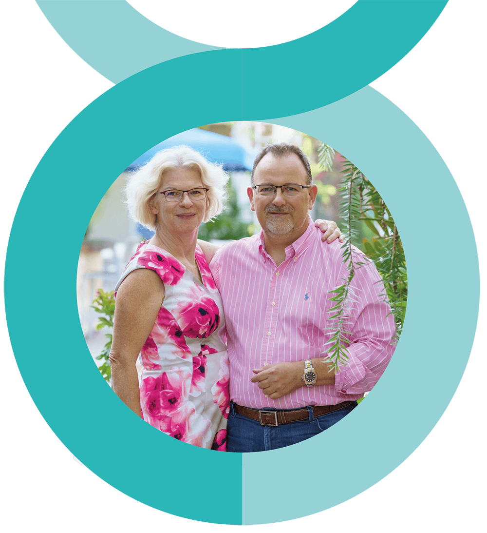 sara-and-andrew A man and woman stand closely together, smiling at the camera. The woman wears a pink floral dress, and the man wears a pink striped shirt and glasses. They are outdoors with greenery in the background.