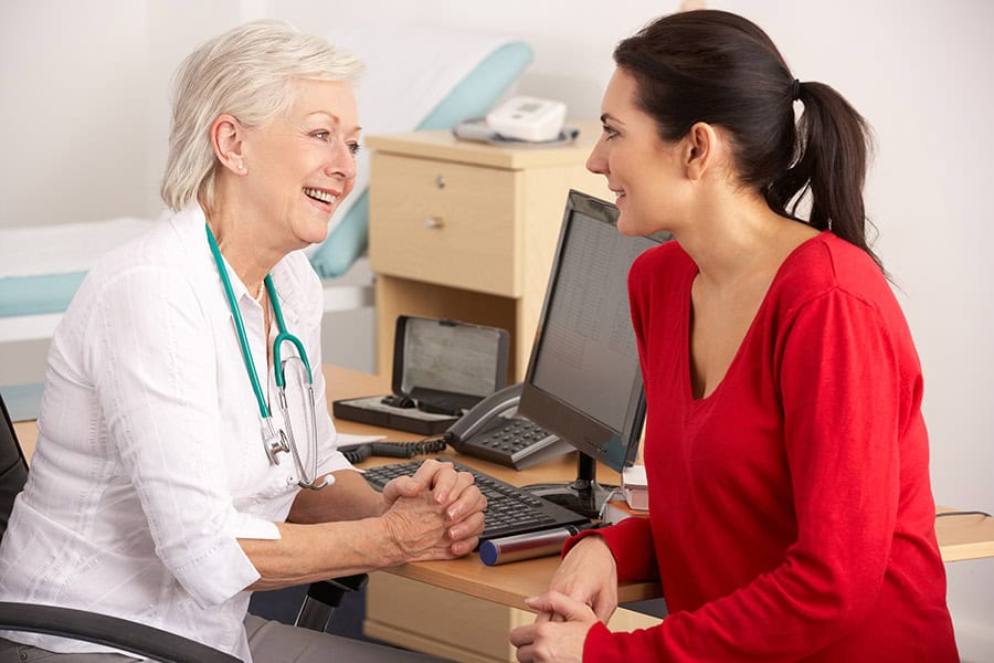 A doctor in a white coat speaks with a patient in a red sweater at a desk in a medical office, both appearing engaged in conversation.