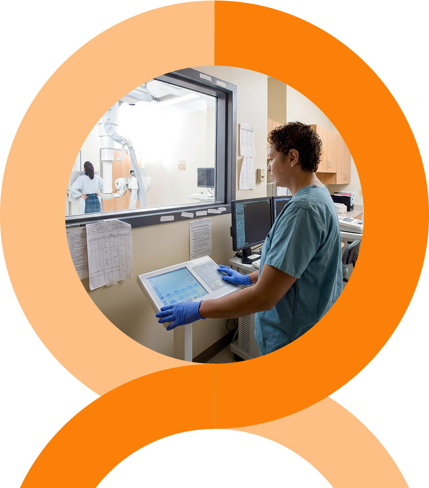 A man and woman stand closely together, smiling at the camera. The woman wears a pink floral dress, and the man wears a pink striped shirt and glasses. They are outdoors with greenery in the background.