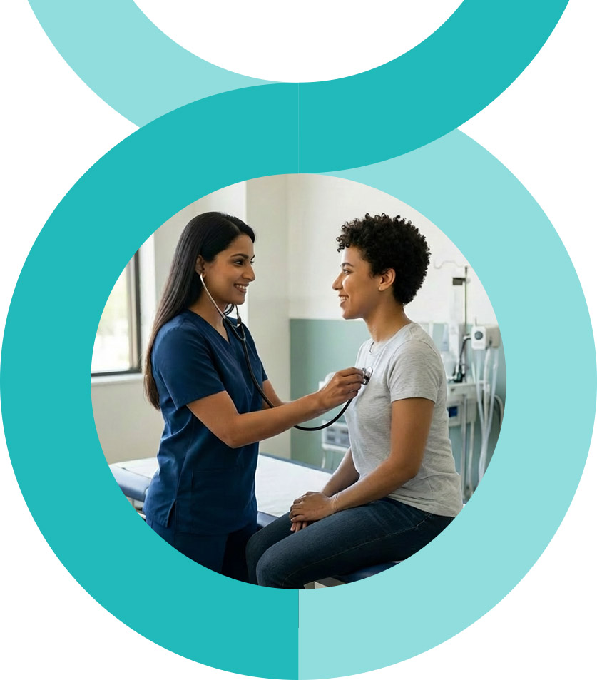 A man and woman stand closely together, smiling at the camera. The woman wears a pink floral dress, and the man wears a pink striped shirt and glasses. They are outdoors with greenery in the background.