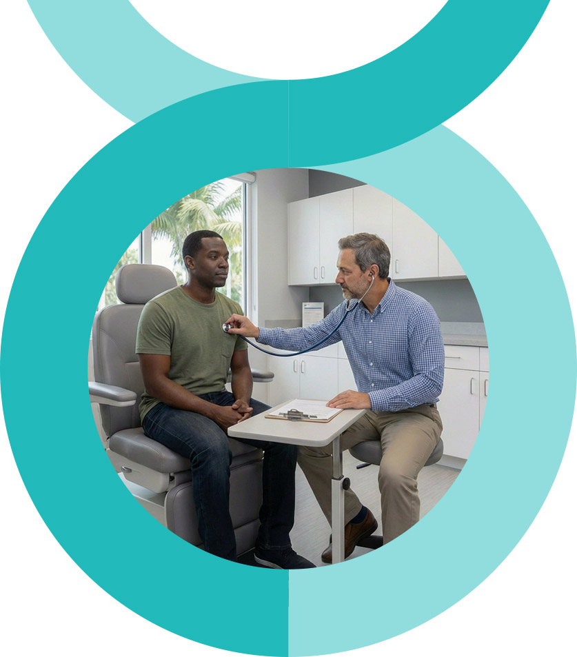 A man and woman stand closely together, smiling at the camera. The woman wears a pink floral dress, and the man wears a pink striped shirt and glasses. They are outdoors with greenery in the background.