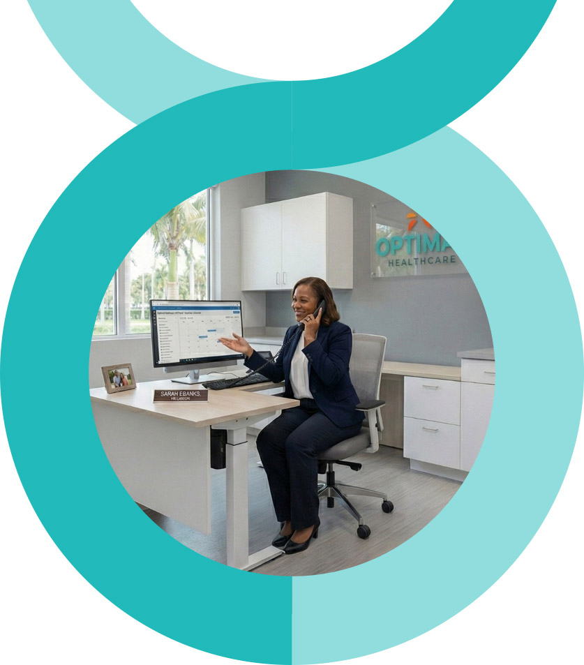 A man and woman stand closely together, smiling at the camera. The woman wears a pink floral dress, and the man wears a pink striped shirt and glasses. They are outdoors with greenery in the background.