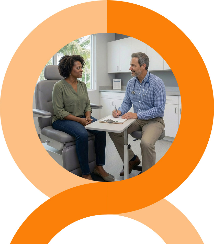 A man and woman stand closely together, smiling at the camera. The woman wears a pink floral dress, and the man wears a pink striped shirt and glasses. They are outdoors with greenery in the background.