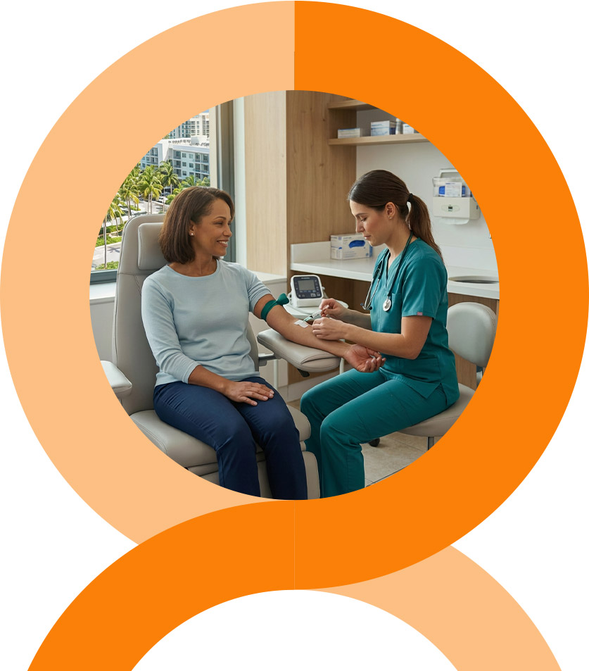 A man and woman stand closely together, smiling at the camera. The woman wears a pink floral dress, and the man wears a pink striped shirt and glasses. They are outdoors with greenery in the background.