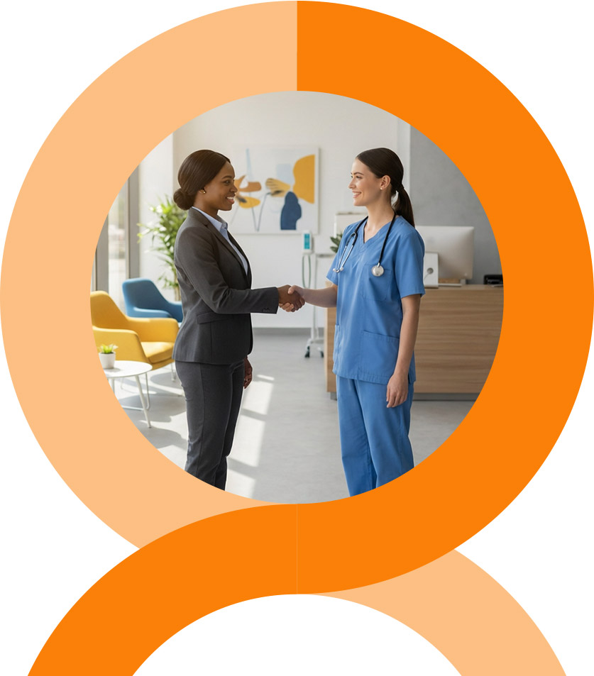 A man and woman stand closely together, smiling at the camera. The woman wears a pink floral dress, and the man wears a pink striped shirt and glasses. They are outdoors with greenery in the background.