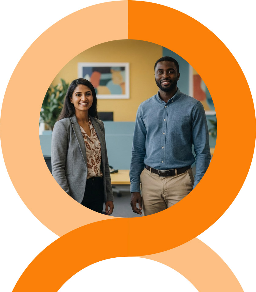 A man and woman stand closely together, smiling at the camera. The woman wears a pink floral dress, and the man wears a pink striped shirt and glasses. They are outdoors with greenery in the background.