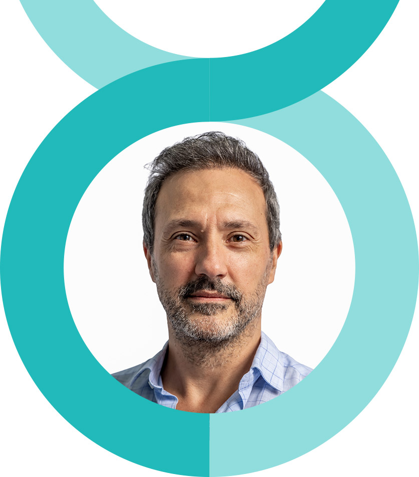 dr-paulo-beco A man and woman stand closely together, smiling at the camera. The woman wears a pink floral dress, and the man wears a pink striped shirt and glasses. They are outdoors with greenery in the background.